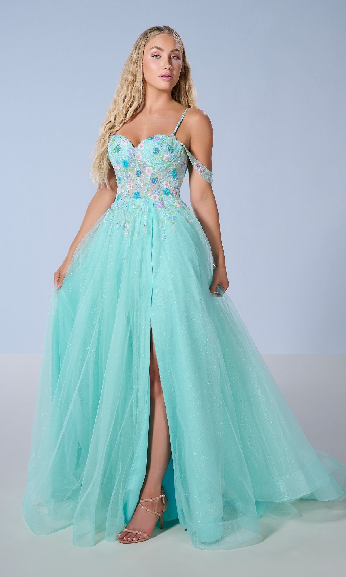 A woman in the Tiffany 16251 long prom dress—light blue, floor-length, a-line with floral beaded appliqué, spaghetti straps, and a high front slit—stands against a plain background.